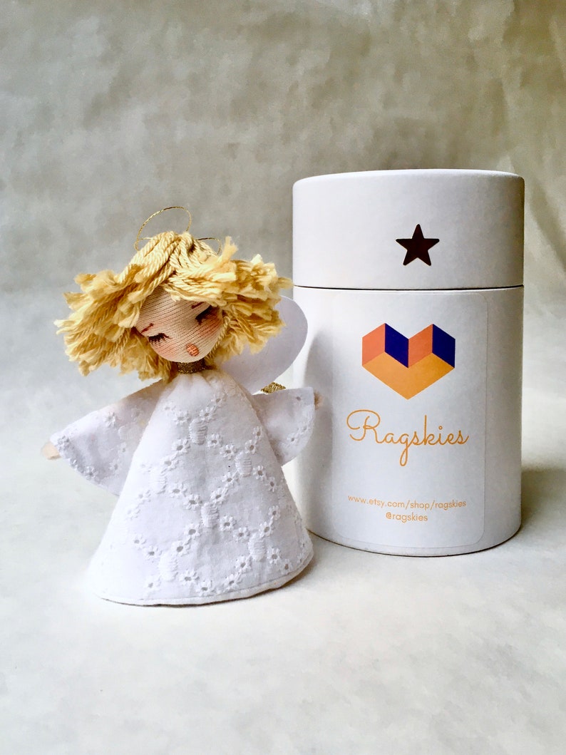 May include: A white fabric angel ornament with a blonde yarn hair and a white lace dress. The angel is packaged in a white cylinder box with a star and a heart logo that says "Ragskies" and includes the website address "www.etsy.com/shop/ragaskies" and the social media handle "@ragaskies".