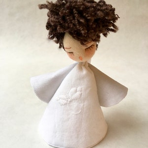 May include: A handmade white fabric angel doll with brown curly hair and closed eyes. The doll is wearing a white dress with a small embroidered flower design.