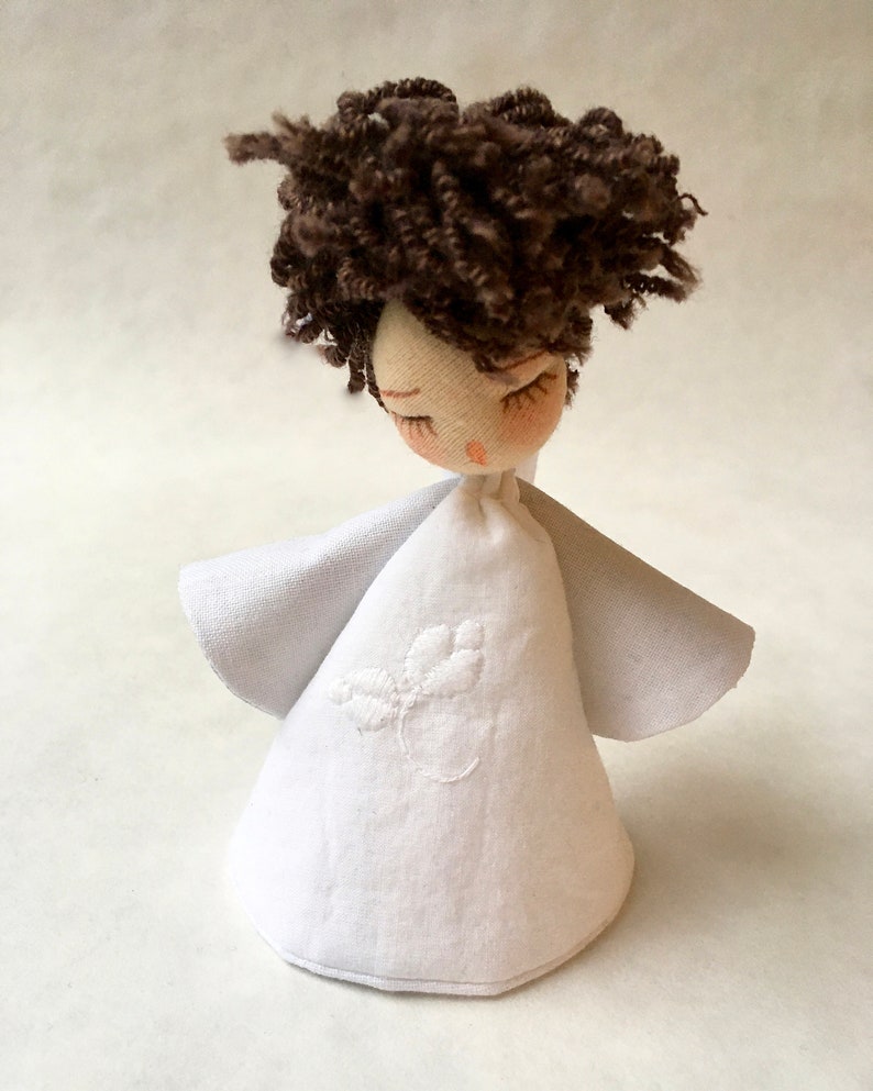 May include: A handmade white fabric angel doll with brown curly hair and closed eyes. The doll is wearing a white dress with a small embroidered flower design.