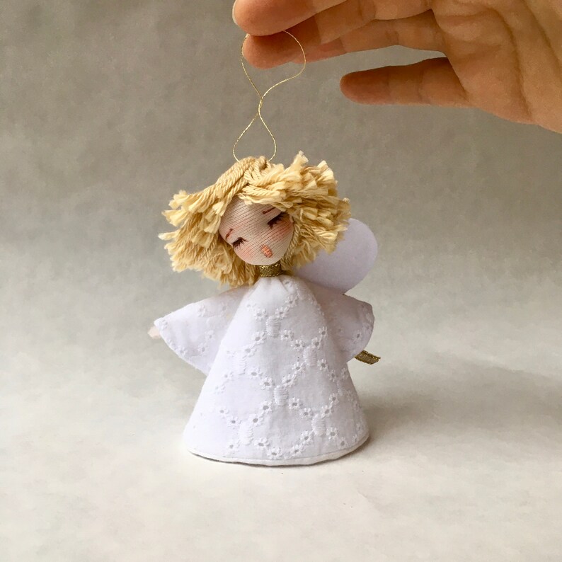 May include: A white fabric angel ornament with a gold halo and blonde yarn hair. The angel has a white dress with a lace pattern and a small white wing.