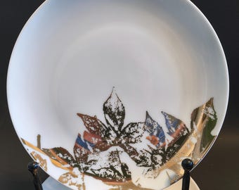May include: White ceramic plates with a leaf design in shades of brown, green, and gold. The plates are displayed on a black metal stand. The leaf design is repeated on the smaller plates below.