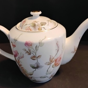May include: White porcelain teapot with a lid, handle, and spout. The teapot is decorated with pink and peach roses and green leaves. Gold trim accents the rim, lid, and spout. The teapot is designed for serving tea.