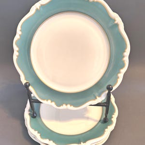 May include: A stack of decorative plates with a teal border and a white center. The plates have a scalloped edge with gold trim. The plates are stacked on a black metal stand.
