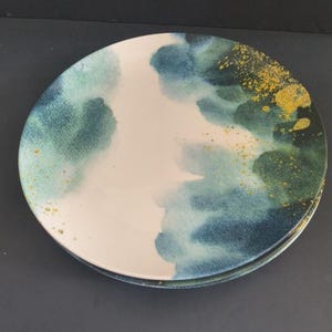 May include: A stack of two round ceramic plates. The plates feature a watercolor design with shades of blue and green, accented with gold flecks. The center of the plates is a creamy white color.