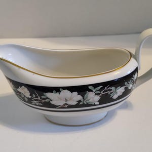 May include: White porcelain gravy boat with a black and gold trim and a floral design featuring white flowers. The boat has a handle and a rounded base.