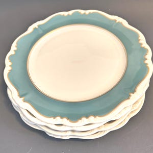 May include: A stack of four ceramic plates. Each plate has a white center, a teal blue border, and a gold trim. The edges of the plates have a scalloped design. The plates are stacked on a gray surface.