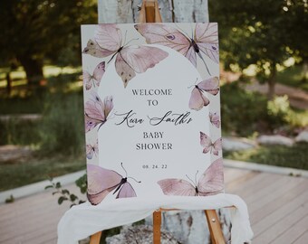 May include: A baby shower welcome sign on an easel. The sign features a soft, watercolor design of butterflies in shades of pink and purple. Text reads "Welcome to Kara Smith's Baby Shower" with the date 08.24.22.