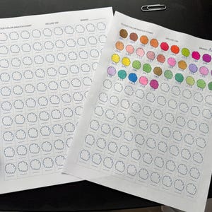 May include: Two white paper color swatch charts labeled "Cloud Color Swatch Chart Deluxe 100". One chart is blank, the other has cloud-shaped color samples filled in with various colors. A silver paper clip rests on the surface.