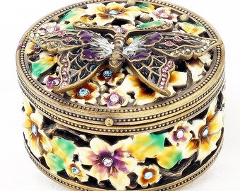 Puede incluir: Caja de baratijas redonda decorada con una mariposa en la tapa. La mariposa tiene alas moradas y negras con gemas rosas. La caja está decorada con diseños florales y pequeñas gemas. La caja es de metal.