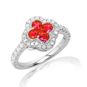 May include: A silver ring featuring a four-leaf clover design. The center of the clover is composed of four red gemstones, surrounded by a halo of small, clear gemstones. The ring band is also adorned with small, clear gemstones.