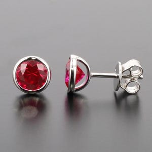 May include: A pair of stud earrings featuring round, faceted red gemstones set in a silver bezel. One earring is shown from the side, displaying the post and butterfly backing. The earrings are displayed on a dark gray surface.