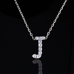 May include: A silver necklace with a pendant in the shape of the letter "J". The pendant and chain are silver. The letter "J" is encrusted with small, clear gemstones. The necklace is displayed against a black background.