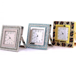 Puede incluir: Tres relojes despertadores de forma cuadrada con un diseño de cristal. Los relojes tienen una esfera blanca con agujas negras y números negros. Los relojes son plateados, azules y dorados con un diseño de estampado de leopardo.