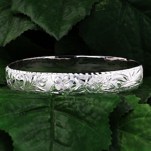 May include: A silver bangle bracelet with a floral design. The bracelet is on a green leafy background.