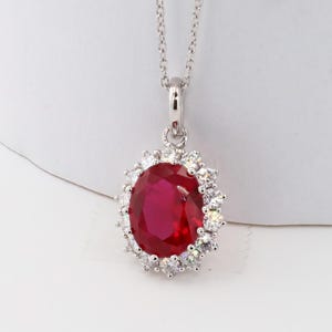 May include: A silver pendant necklace featuring a large, oval-cut ruby gemstone surrounded by a halo of small, clear gemstones. The ruby is a deep red color, and the necklace hangs from a delicate silver chain.