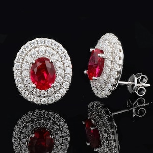 May include: A pair of stud earrings featuring a vibrant red oval gemstone surrounded by a halo of smaller, sparkling white stones. The earrings are set in a silver metal and reflect on a black surface.