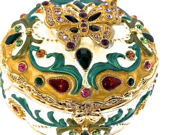 Puede incluir: Una caja redonda de baratijas blanca con un borde dorado y un diseño de mariposa. La mariposa está hecha de oro y tiene piedras de imitación para las alas. La caja está decorada con acentos verdes, dorados y rojos.