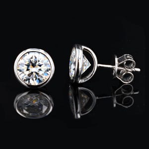 May include: Two silver stud earrings with a large, round, clear gemstone. The gemstones are set in a bezel setting. The earrings are displayed on a black surface, with reflections visible.
