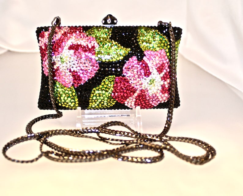 May include: A black evening bag with a chain strap. The bag is covered in rhinestones and features a floral design with pink, green, and clear rhinestones.
