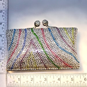May include: A small, silver-toned clutch purse covered in colorful rhinestones. The rhinestones are arranged in a pattern of diagonal stripes, creating a rainbow effect.