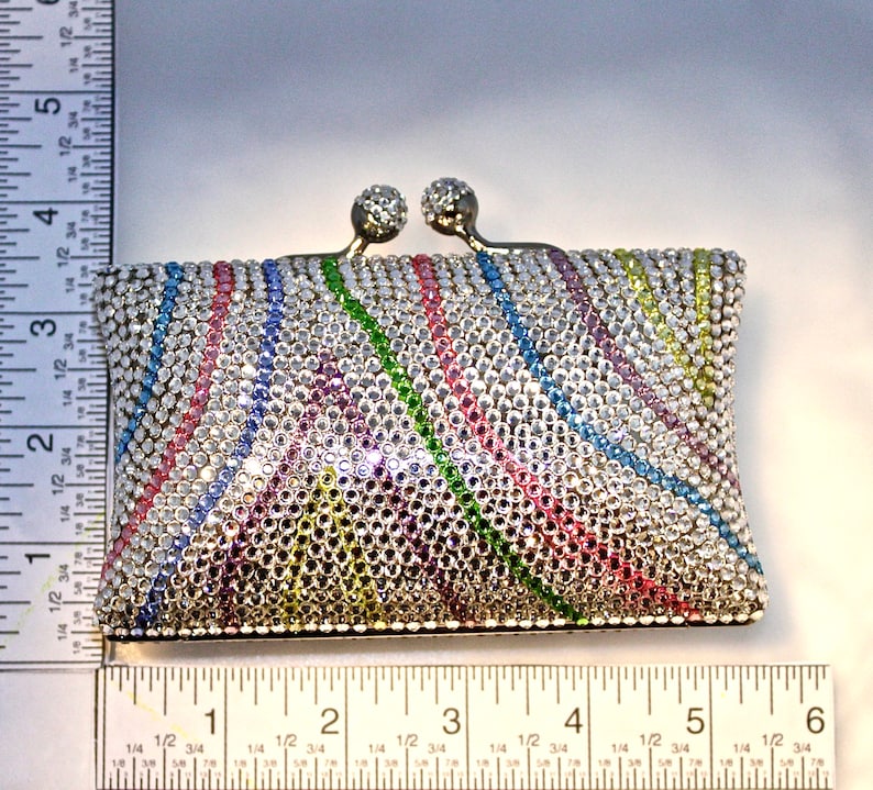 May include: A small, silver-toned clutch purse covered in colorful rhinestones. The rhinestones are arranged in a pattern of diagonal stripes, creating a rainbow effect.