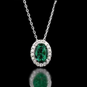 Puede incluir: Un collar de plata con un colgante que presenta una gran esmeralda ovalada rodeada por un halo de pequeños diamantes brillantes. El colgante cuelga de una delicada cadena de plata sobre un fondo negro.