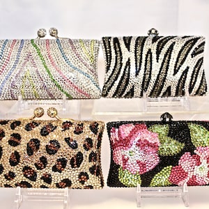 May include: Four different rhinestone clutch purses. The purses are displayed on a white surface with clear acrylic stands. The purses are decorated with different patterns, including zebra stripes, leopard print, and floral designs.