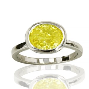 Könnte beinhalten: Ein silberner Ring mit einem großen, ovalen gelben Edelstein. Der Edelstein ist in einer Zargenfassung gefasst, und das Band ist glatt und poliert. Der Ring wird auf einer reflektierenden Oberfläche gezeigt.