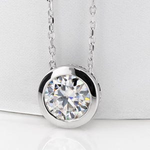 May include: A silver pendant necklace with a large, round, clear gemstone set in a bezel. The gemstone has multiple facets, creating a sparkling effect. The chain is delicate and silver. The necklace is shown against a white backdrop.