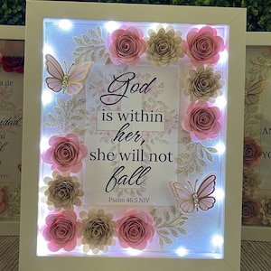 May include: A white framed art piece with the text "God is within her, she will not fall" surrounded by pink and gold paper flowers and butterflies. The frame has small lights.