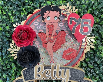 May include: A red, white, and black cake topper featuring Betty Boop in a red dress with a red heart that says "75" on it. The topper also has a black banner with the word "Betty" on it.