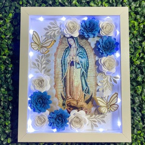 May include: A white framed picture with a light inside, featuring a religious image of the Virgin Mary with blue and white paper flowers surrounding the image. The flowers are arranged in a circle around the image.