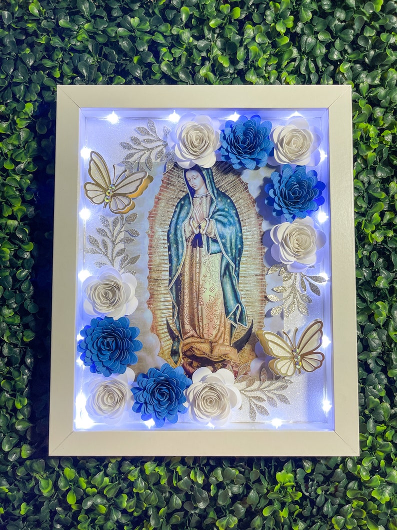 May include: A white framed picture with a light inside, featuring a religious image of the Virgin Mary with blue and white paper flowers surrounding the image. The flowers are arranged in a circle around the image.