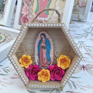 Puede incluir: Una caja de sombra hexagonal de madera con una imagen de Nuestra Se&ntilde;ora de Guadalupe. El marco est&aacute; adornado con pedrer&iacute;a y flores de papel en tonos amarillos y fucsia. Se adjunta un asa de cuerda de yute para colgar.