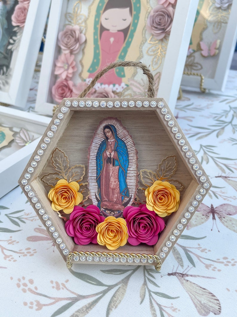 Puede incluir: Una caja de sombra hexagonal de madera con una imagen de Nuestra Se&ntilde;ora de Guadalupe. El marco est&aacute; adornado con pedrer&iacute;a y flores de papel en tonos amarillos y fucsia. Se adjunta un asa de cuerda de yute para colgar.