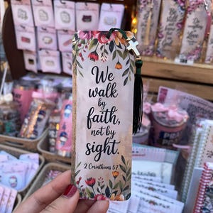 May include: A floral bookmark with the text "We walk by faith, not by sight. 2 Corinthians 5:7." The bookmark has a green tassel and a small cross charm. The design includes red, pink, and yellow flowers.