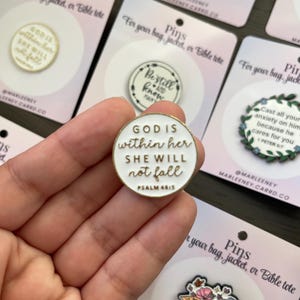 May include: A selection of enamel pins with motivational quotes. The pins are white with gold edging and feature phrases such as "God is within her, she will not fall." The pins are displayed on cards that read "Pins for your bag, jacket, or Bible tote."