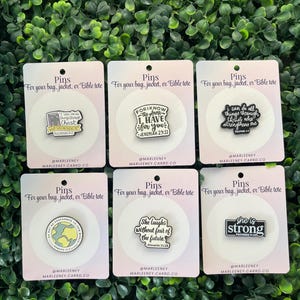 May include: Six enamel pins with inspirational quotes on white cards. The pins feature phrases like "I can do all things through Christ" and "She laughs without fear of the future." Each pin is designed for bags, jackets, or Bible totes.