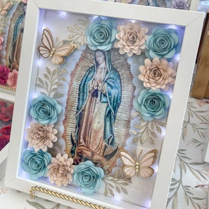 May include: A white frame with a light blue, pink, and gold floral design. The frame contains a picture of the Virgin Mary and a butterfly. The frame is lit with LED lights.