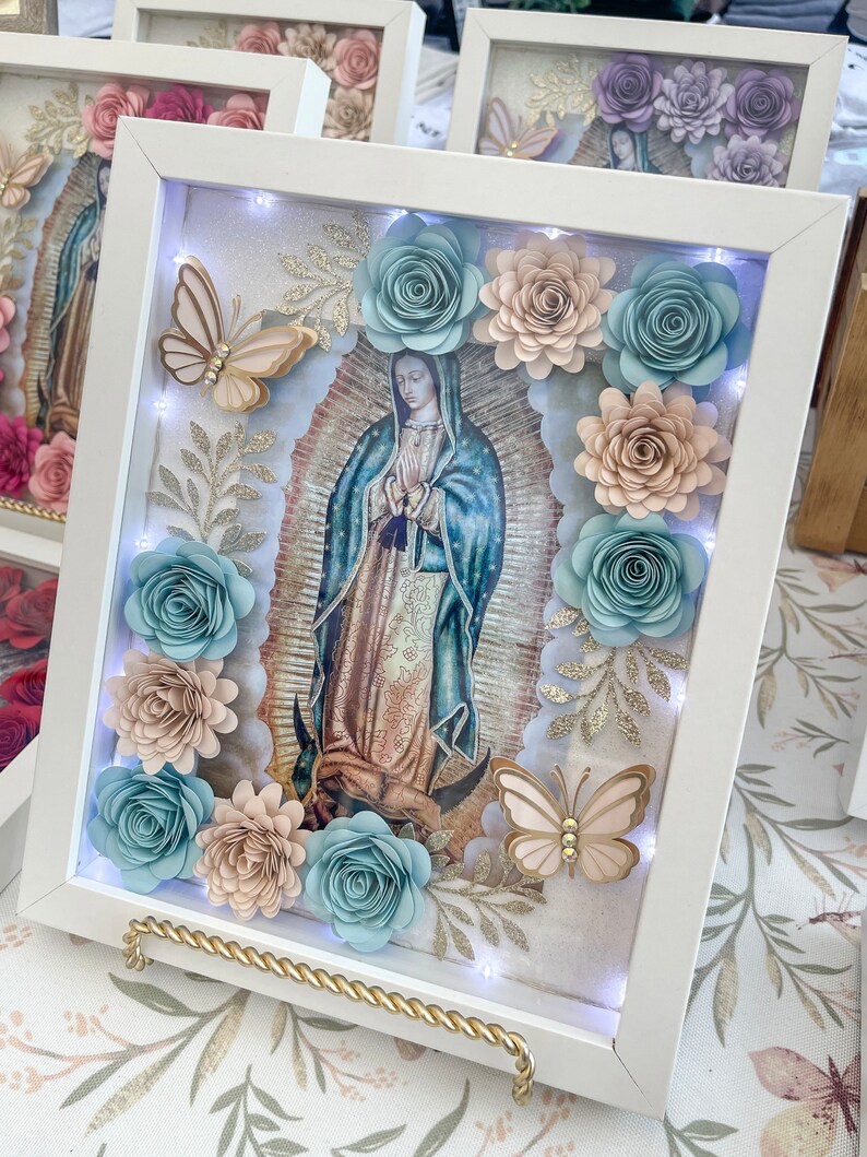 May include: A white frame with a light blue, pink, and gold floral design. The frame contains a picture of the Virgin Mary and a butterfly. The frame is lit with LED lights.