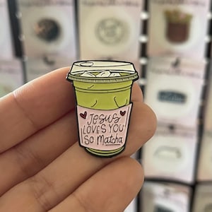 May include: A green matcha latte enamel pin with a clear lid. The pin features a pink sleeve with the text "Jesus Loves You So Matcha" in a handwritten font. Two small hearts are on the sleeve. The pin is held in a hand.