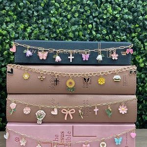 May include: Four gold-toned metal chains with various enamel charms draped across four stacked books. Charms include flowers, butterflies, bees, crosses, hearts, bows, cacti, and cupcakes in pink, yellow, green, white, and blue colors.