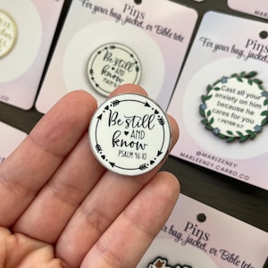 May include: A round enamel pin with the words "Be still and know" and Psalm 46:10 in black on a white background. The pin has a black arrow and dot design around the edge. Other pins are visible in the background.
