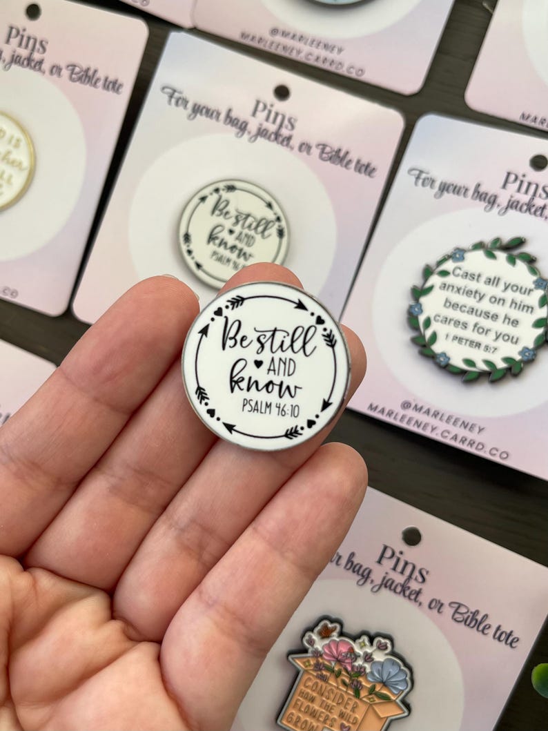 May include: A round enamel pin with the words "Be still and know" and Psalm 46:10 in black on a white background. The pin has a black arrow and dot design around the edge. Other pins are visible in the background.