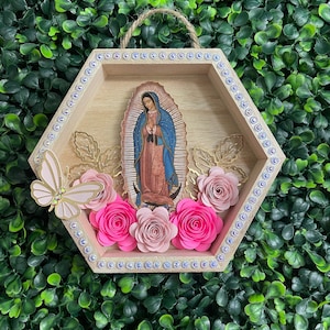 Puede incluir: Un colgante de pared hexagonal de madera con una imagen de Nuestra Se&ntilde;ora de Guadalupe. El marco est&aacute; decorado con pedrer&iacute;a y flores de papel en tonos rosas. Una mariposa y hojas doradas a&ntilde;aden detalles.