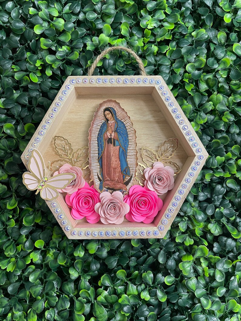 Puede incluir: Un colgante de pared hexagonal de madera con una imagen de Nuestra Se&ntilde;ora de Guadalupe. El marco est&aacute; decorado con pedrer&iacute;a y flores de papel en tonos rosas. Una mariposa y hojas doradas a&ntilde;aden detalles.