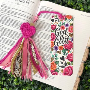 May include: A decorative bookmark with a pink heart, tassels, and the word "Blessed." The bookmark features a floral design with the words "God is Good." The bookmark is placed in an open book.
