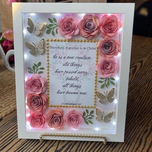May include: A white framed shadow box featuring pink paper roses, glitter butterflies, and the Bible verse 2 Corinthians 5:17. The frame has small white lights and a gold rope trim, creating a decorative and inspirational piece.
