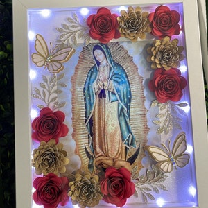 May include: A white frame with a light inside, featuring a picture of the Virgin Mary. The frame is decorated with red and gold paper flowers and butterflies.