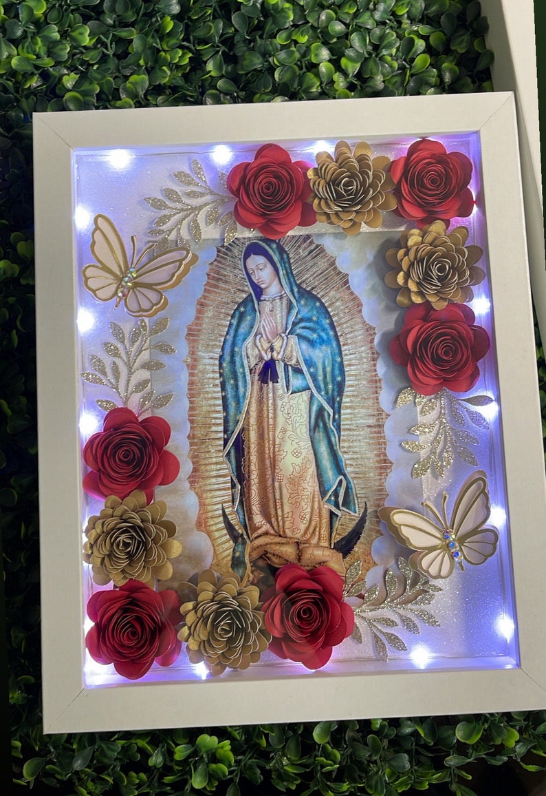 May include: A white frame with a light inside, featuring a picture of the Virgin Mary. The frame is decorated with red and gold paper flowers and butterflies.